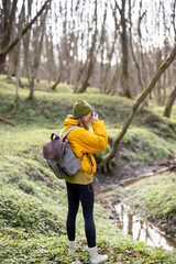 Woman in hiking clothes with backpack takes picture of green spring forest. Enjoys of purity and freshness of nature. Travel alone.