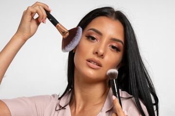 Beautiful long black-haired young woman puts makeup with brush on her face in beauty studio