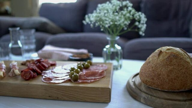 Panning footage across a table set with food and drink. Contemporary home interior setting with natural daylight. Meat platter, sharing snack foods including rustic sourdough, cold meats and olives