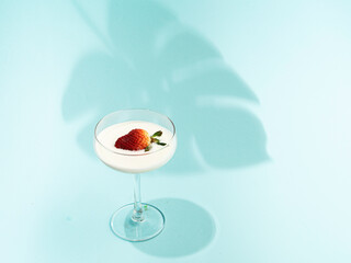 Champagne glass, creamy  fruit yogurt inside, decorated with fresh strawberry, mint blue background, tropical leaf shadow