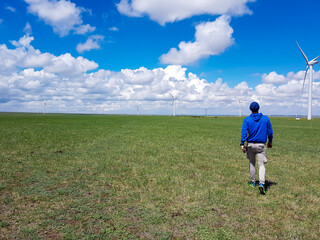 Naklejka na ściany i meble A man walking towards wind turbines build on a vast pasture in Xilinhot in Inner Mongolia. Natural resources energy. Clean energy. Endless grassland. Blue sky with white, thick clouds. Endless horizon
