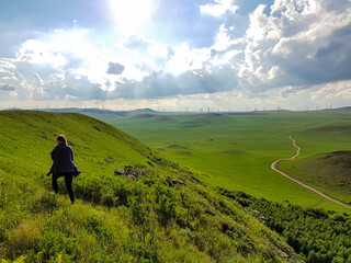 A woman walking on a green hill in Xilinhot, Inner Mongolia. Endless grassland with a few wind turbines in the back. Blue sky with thick, white clouds. A small gravel road through the pasture. Freedom