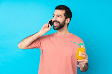 Young man over holding a cocktail over isolated blue background keeping a conversation with the mobile phone