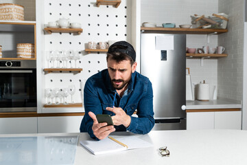 Fototapeta na wymiar Young freelance business man in casual clothing standing at his kitchen at his home and checking the plans and statistic on his cell phone application, work at home concept