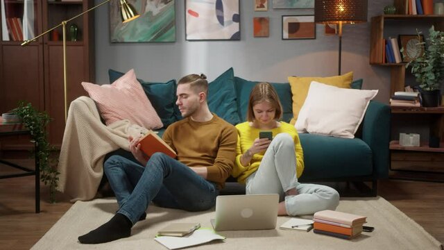 Loving students couple indoors at home reading book, preparing for exams, woman using smartphone. Young family, man read homework, remote learning during coronavirus lockdown.