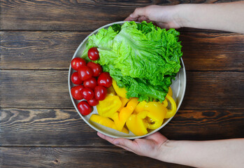 child's hands holding a ceramic plate with fresh vegetables, a healthy snack and diet lettuce leaves, cherry tomatoes and bell peppers on a wooden table top view