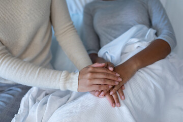 Midsection of mixed race mother comforting sick daughter, sitting on hospital bed holding her hands