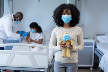 Mixed race woman in face mask holding teddy in hospital doctor giving covid vaccine to sick daughter