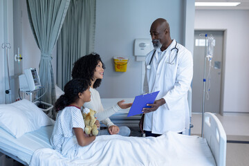 Obraz premium African american male doctor talking with mixed race mother and her sick daughter, in hospital bed