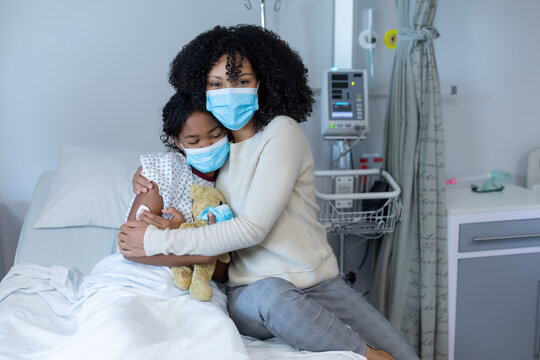 Mixed race mother hugging sick daughter in hospital bed holding teddy bear, all wearing face masks