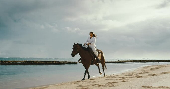 Beautiful young woman horseback riding on the beach, strong and powerful horse galloping in the sand, cinematic slow motion