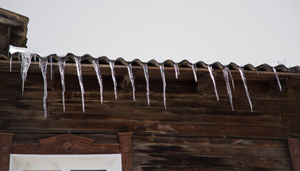 Icicles hang from the roof of a wooden house on a frosty, cloudy day. The slate roof is against the gray sky. Abnormal weather concept. New Year's and Christmas is over.