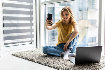 Beautiful girl in casual clothes is doing selfie using a smart phone and smiling while working with a laptop on the floor at home