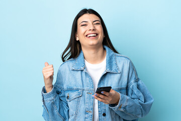 Young brunette girl over isolated blue background with phone in victory position