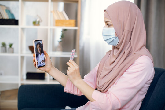 Muslim sick woman in hijab and protective mask, holding cell phone in one hand and pills in another one. African woman doctor therapist on video conference call online, helps her female patient.