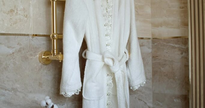 White textile bath robe with floral patterns and shower towel are haging on the gold colered towel warmer in the corner of bathroom. The walls are made of brown marbled tiles