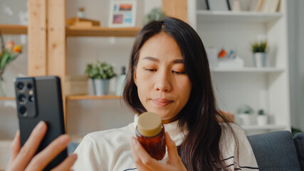Sick young Asian woman hold medicine sit on couch video call with phone consult with doctor at home. Girl take medicine after doctor order, quarantine at home, Social distancing coronavirus concept.