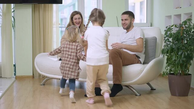 Cheerful Daughters Running In Slow Motion To Couple Of Happy Parents Sitting On Couch In Living Room. Wide Shot Of Joyful Caucasian Young Family Resting Together On Weekends. Unity Concept