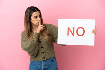 Young caucasian woman isolated on pink background holding a placard with text NO doing silence gesture