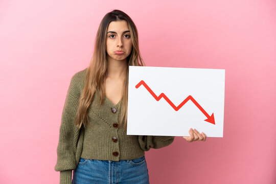 Young caucasian woman isolated on pink background holding a sign with a decreasing statistics arrow symbol with sad expression