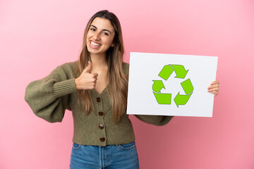 Young caucasian woman isolated on pink background holding a placard with recycle icon with thumb up