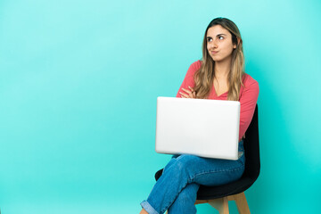 Young caucasian woman sitting on a chair with her pc isolated on blue background making doubts gesture while lifting the shoulders