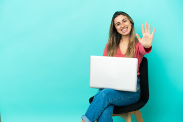 Young caucasian woman sitting on a chair with her pc isolated on blue background counting five with fingers