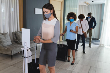 Diverse businesswomen and businessmen standing in line to disinfect hands in hotel lobby