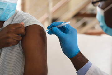 African american senior male doctor injecting covid-19 vaccine into african american man at home