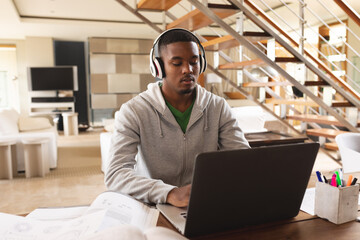 African american young man wearing headphones using laptop while studying at home