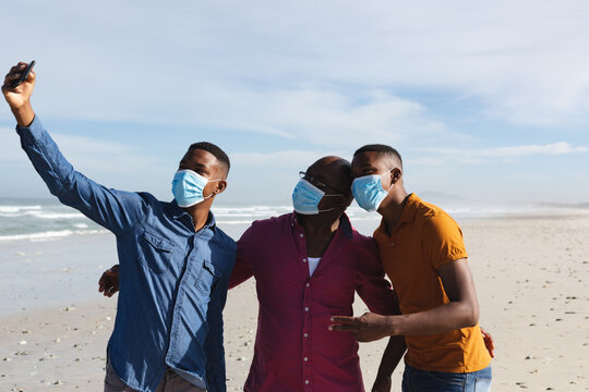 African american father and his two sons wearing face masks taking a selfie together at the beach