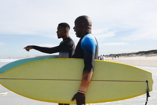 African american father and son with surfboards walking towards the waves at the beach