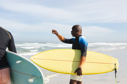 African american senior man with surfboard pointing towards the waves at the beach