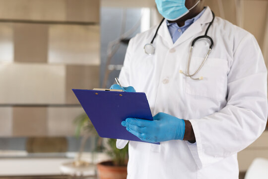 Mid section of african american senior male doctor wearing face mask writing on clipboard at home