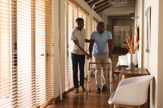 African american young man helping his father to walk with walking frame at home