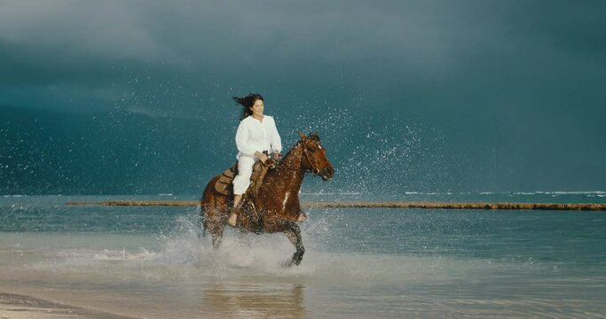 Beautiful young woman horseback riding on the beach, strong and powerful horse splashing in the water, cinematic slow motion