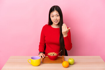 Young Chinese girl  having breakfast in a table inviting to come with hand. Happy that you came