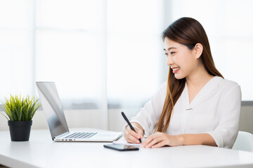 Young asian beautiful business woman working with laptop sitting at home and note on book. Smiling charming happy young female doing homework meeting conference with team at home.