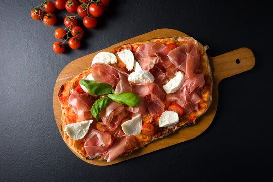 Pinsa with San Daniele raw ham, mozzarella, cherry tomatoes, basil on a cutting board near a bunch of cherry tomatoes