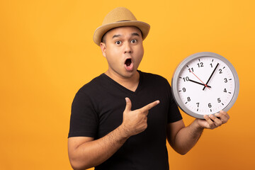 Young asian man wearing hat black shirt holding Big clock standing on isolated yellow background.