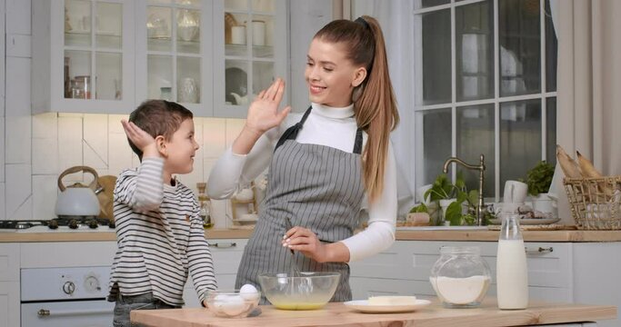 Young woman preparing dough with her little son, giving high five to each other and laughing to camera at kitchen