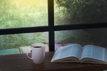 morning fresh black hot coffee in white cup with opened book on wood table with blurred black windows frame and green garden home's yard in background