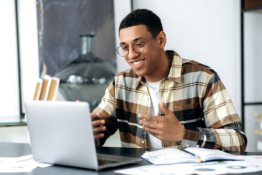 Attractive friendly mixed race latino freelancer guy with glasses, sits at a desk, uses laptop, have a video call, communicate with colleagues or friends by video conference, gesturing hands, smiling