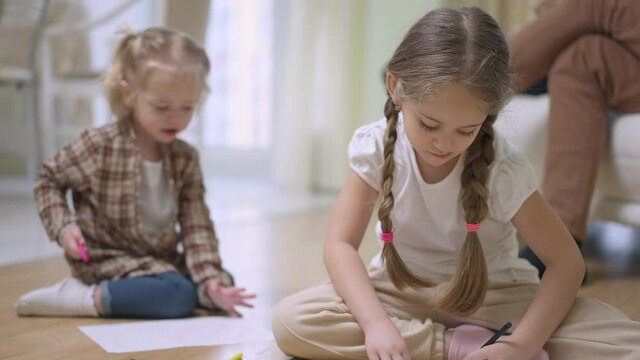 Portrait of concentrated charming Caucasian girl with pigtails painting sitting on floor with blurred baby sister drawing at background. Positive relaxed children enjoying weekend at home indoors