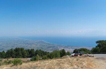 Obraz premium View from the mountain to the valley and the blue sea. A lonely truck is standing on a hill, and just below is an asphalt highway. Blue sky with white cumulus clouds. Summer time. Horizontal photo. 