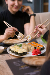 Two women sit at the table and hold sushi with chopsticks over a plate of sushi