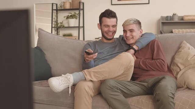 Lockdown of pair of young cheerful Caucasian men wearing casual clothes sitting in embrace on sofa at home and watching funny show on TV