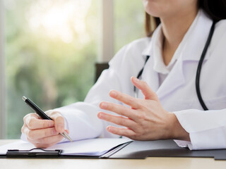 A close-up of the hand of a female doctor or nurse is holding a pen and using another hand to explain information or something with a blurred background.