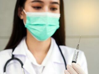 A female doctor or nurse with a surgical mask and stethoscope in blurred is holding a syringe, close-up on a syringe in her hand.