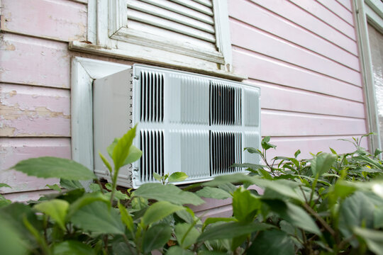 An old clunky white air conditioning system with metal grills installed on the outside of a pink panelled wooden home with a green garden hedge in the foreground. Shot in Barbados. Heat wave concept.